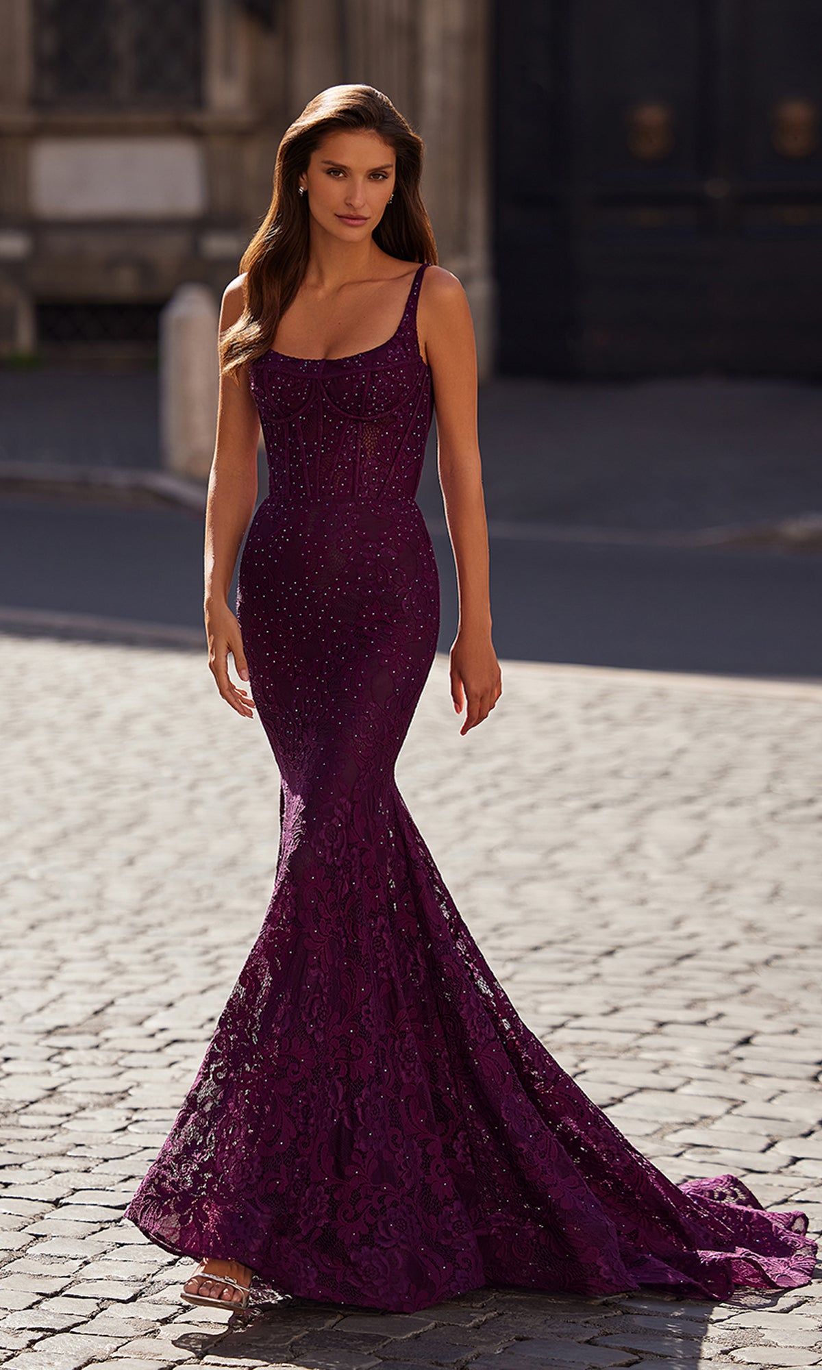 A woman wearing the Long Lace Mermaid Prom Dress: La Femme 32420 with a fitted, floor-length silhouette and flared skirt walks on a cobblestone street—a stunning formal gown ideal for any elegant event.